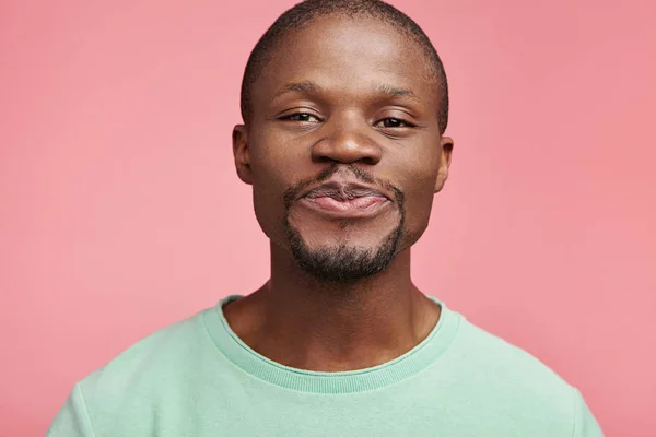 Pleased kind hearted dark skinned male presses lips, looks with dark narrow eyes into camera, isolated over pink background.