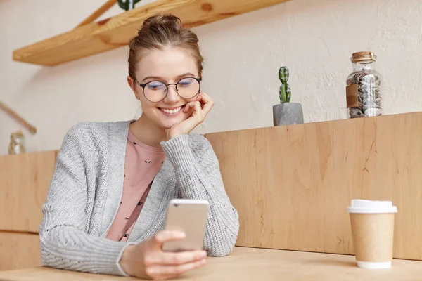 Beautiful young female wears knitted sweater and glasses sits over home interior with smartphone and paper cup of coffee, has happy expression as receives message from lover, invitation for date