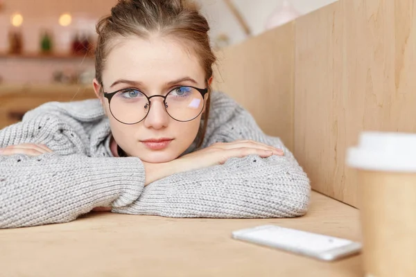 Thoughtful young female wears knitted sweater and round glasses, has autumn depression, being upset and deep in thoughts, sits at wooden table in cafe, tries to escape from all difficulties.