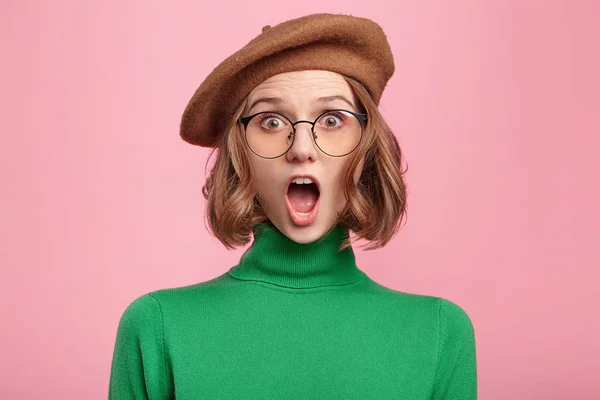 Excited female nerd wears beret and spectacles, looks surprise into camera, shocked. Fearful young pretty woman afraid of something