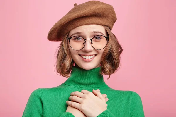 Portrait of smiling positive woman keeps hand on chest, expresses her sympathy. Kind hearted friendly young female shows kindness, wears beret and green turtleneck sweater, has joyful expression.