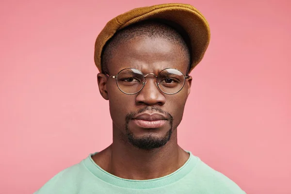 Stern serious dark skinned male looks through spectacles, frowns face in dissatisfaction, displeased as receives very low salary, can`t pay all bills and debts, poses against pink background.