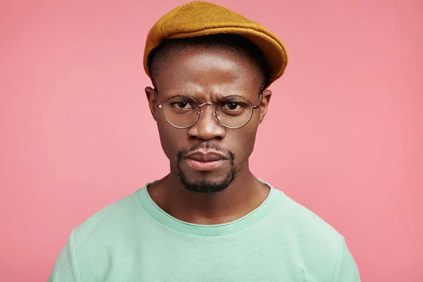 Outraged serious strict male boss looks through glasses, wears fashionable clothes, dissatisfied with results of sales, isolated over pink background. Discontent furious dark skinned young man