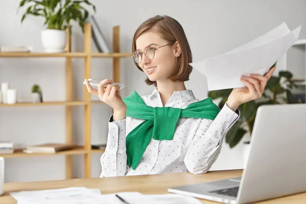 Portrait of talented female entrepreneur involved at working process, holds papers, makes voice call, discuss main issues with colleagues, being always in touch. Woman absorbed in financial issues