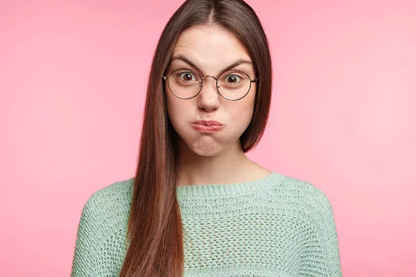 Annoyed young female blowing cheeks in dissatisfaction, raising eyebrows and frowning face on pink background