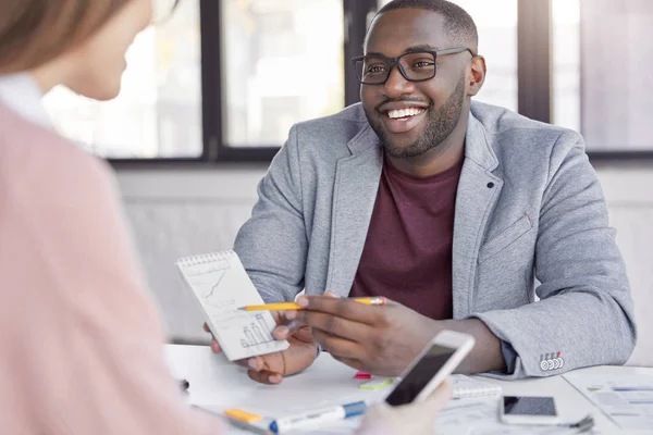 African male in formal clothes discusses details of new project to his assistant, explains charts and diagrams, points at notebook. Two mixed race coworkers talk about bargain during office meeting