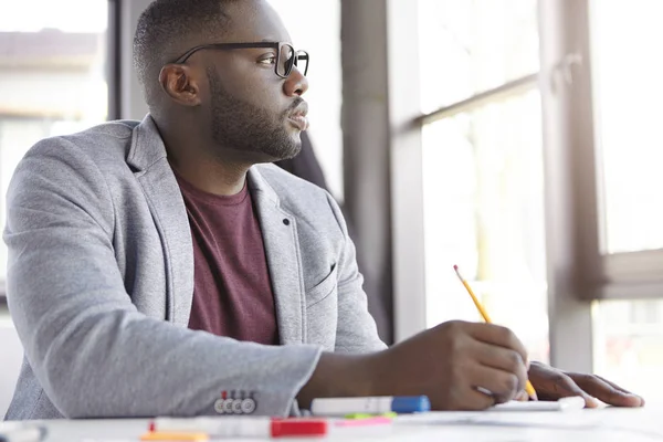 Serious dark skinned plump male businessman, studies important documents, thinks how complete online project, looks pensively aside, generates new ideas, writes down general concepts in notebook