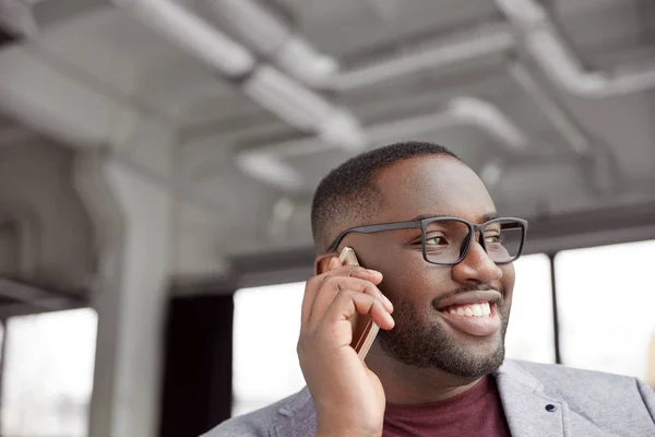 Horizontal shot of successful male with dark skin, wears spectacles, has pleasant smile as talks over smart phone with business partner, looks aside, discuss working schedule together.