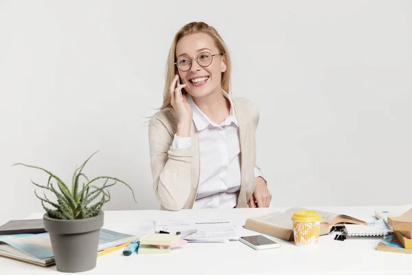 Cute prosperous blonde businesswoman has phone talk, share ideas with partner about new productivity project for company, has good mood, sits at work place. Technology and occupation concept