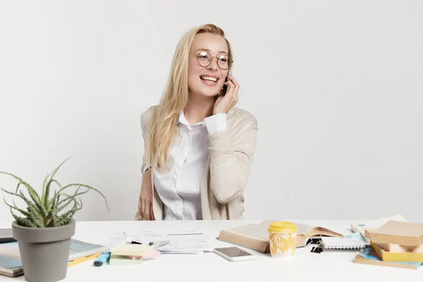 Portrait of female executive manager in eyewear, satisfied with report, shares success to companion or friend over smart phone. Cheerful businesswoman books tickets online, talks with operator