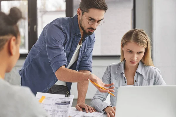 Crew of creative young talented professionals create new startup project, look attentively into screen of laptop computer, analyzes documents. Handsome male tries to present his brilliant ideas