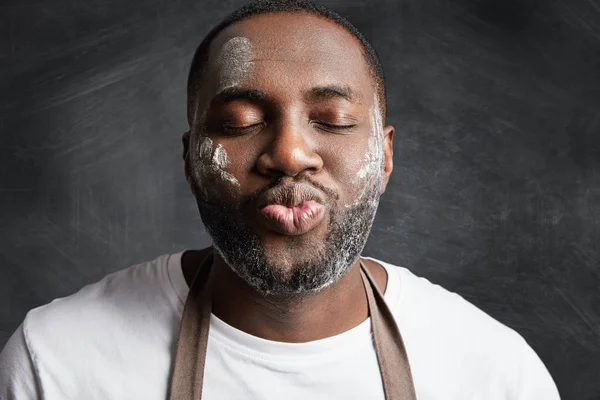 Close up portrait of dark skinned male being satisfied after baking something delicious, rounded lips and closes eyes in enjoyment, has dirty face with flour poses against black chalk background.