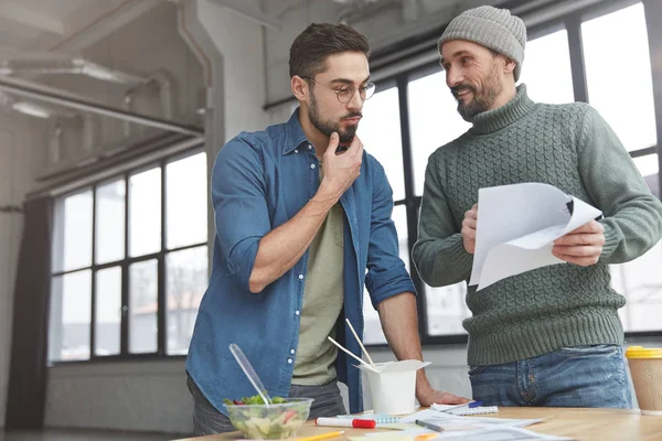 Cheerful mature bearded male teaches young male office worker how to prepare business report, shows on which figures he should pay attention, eat Asian exotic food and fresh vegetable salad