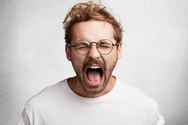 Portrait of emotional crazy male yells loudly, being irritated with someone or something, expresses negative emotions, isolated over white background. Mad stylish man shouts or screams at friend