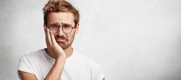 Discontent sorrowful male has terrible toothache, can`t stand pain, being upset, poses against white background with copy space for your advertising content. Upset unhappy man has some troubles