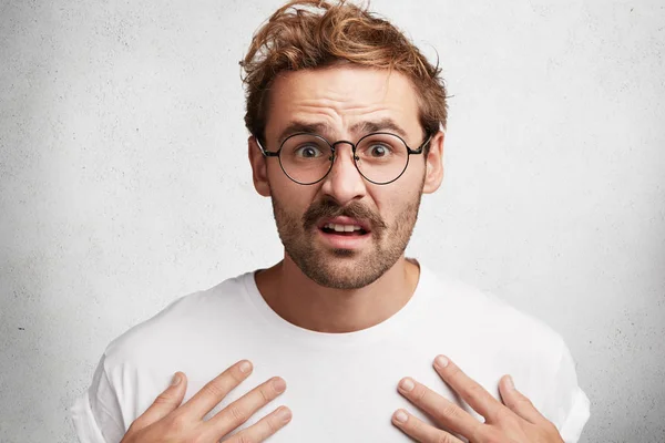 Indignant puzzled young bearded male with discontent expression, points at himself, wears casual white t shirt and spectacles, poses against white concrete wall. People and body language concept