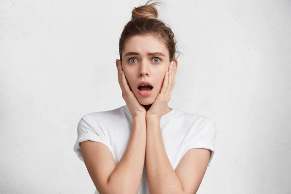 Surprised young pretty female has hair tied in knot, looks with opened mouth, shocked to see something awful, dressed casually, isolated over white background. People, surprise and facial expression