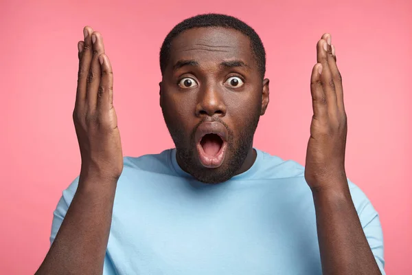 Frightened bugged eyed black man gestures actively, keeps mouth widely opened, afraid of something awful, isolated over pink background. Shocked dark skinned male poses and gestures in studio