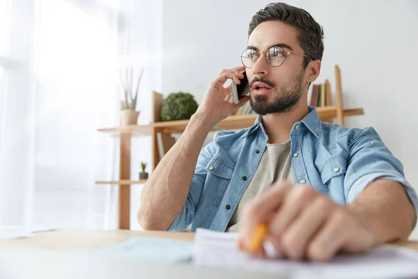 Serious concentrated fashionable male office worker calls to colleague on cellular, has phone talk, try to arrange meeting discuss everything alive. Creative male designer shares idea with companion