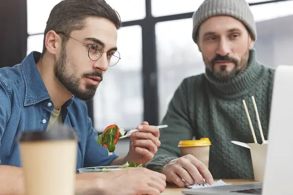 Concentrated serious young male vegan has balanced diet, eats low calorie healthy salad while studies statistics on laptop computer with his companion, collaborate for work, have snack in office