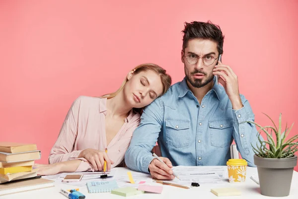 Busy male freelancer writes in documents, has phone conversation while female partner leans at his shoulder, fell asleep, being overworked and fatigue, need rest. People, work, tiredness concept
