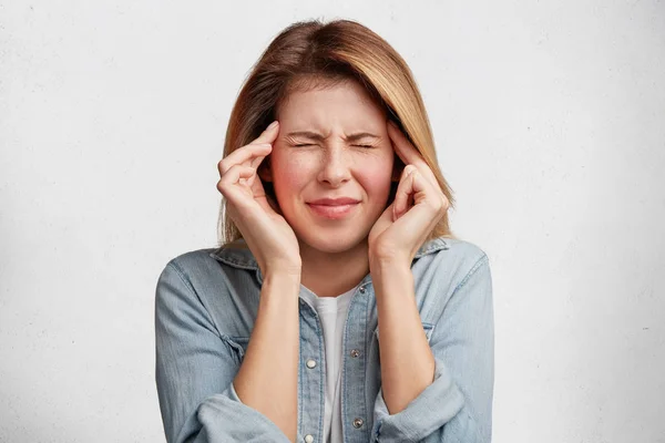 Indoor shot of depressed female model wears denim jacket, keeps finger on temples, suffers from headache, feels pain, being overworked, need rest and calm.