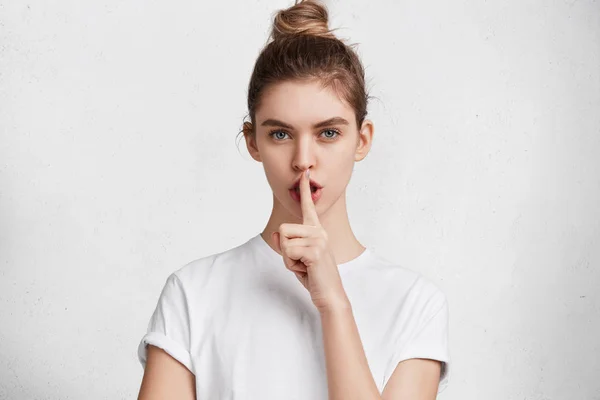 Horizontal shot of beautiful female wears casual white t shirt, shows hush sign, has serious expression, asks best or close friend not to tell secret anybody, hopes for loyalty and silence.