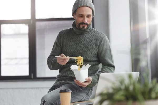 Delightful middle aged male with thick beard and mustache has dinner break in office, eats noddle with chopsticks and watches film on laptop computer, enjoys delicious fast food.