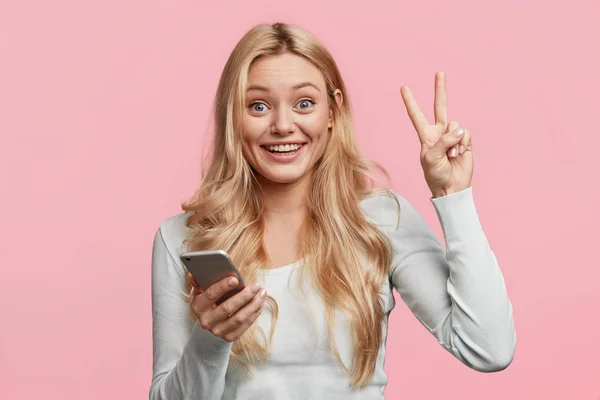 Portrait of pretty young female blogger with peace gesture and smartphone on pink background