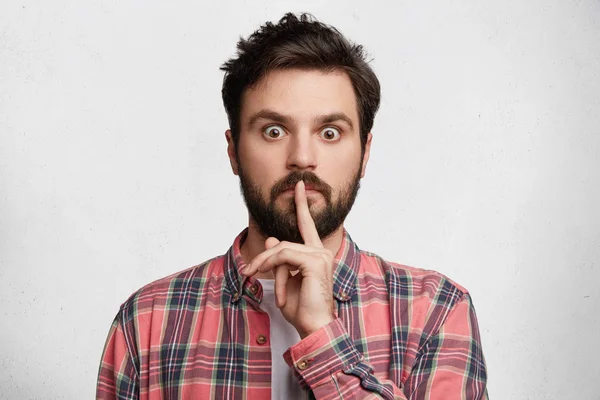 Frightened scared male gives instructions to keep silence, wears checkered shirt, has thick beard and mustache, isolated over white studio background. Fashionable guy shows shush sign, asks be quiet