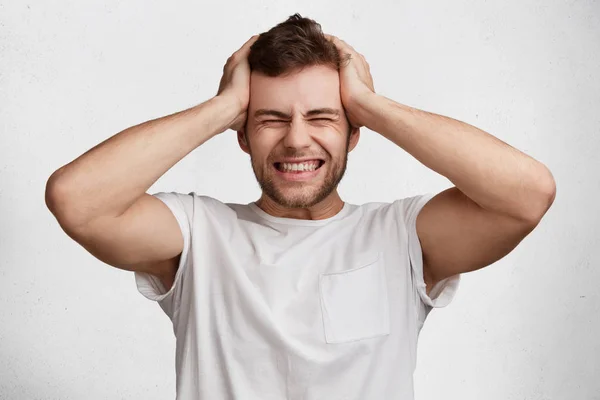 Desperate stressful bearded male clenches teeth and scratches head, being dissatisfied with current situation, isolated over white background. Frustrated European male in despair, has headache