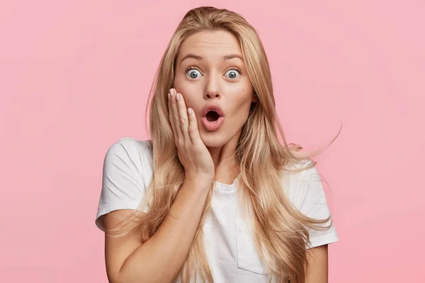 Indoor shot of stupefied shocked blonde woman keeps mouth widely opened, looks in terror at camera, wears casual white t-shirt, isolated over pink background. Attractive female model indoor.