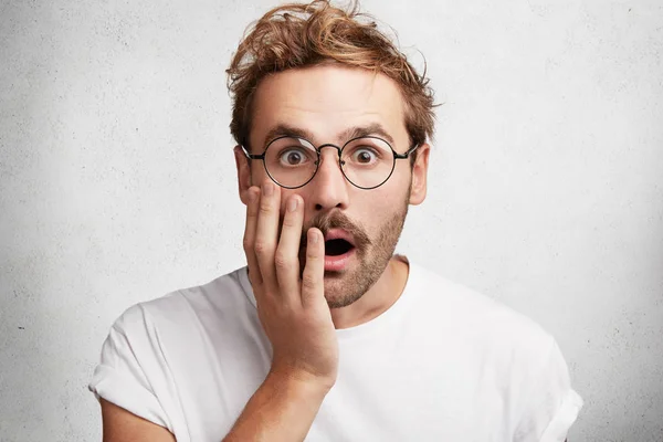 Portrait of shocked frightened bearded young male stares at camera through round spectacles, keeps mouth widely opened, covers mouth with hand, isolated over white concrete background