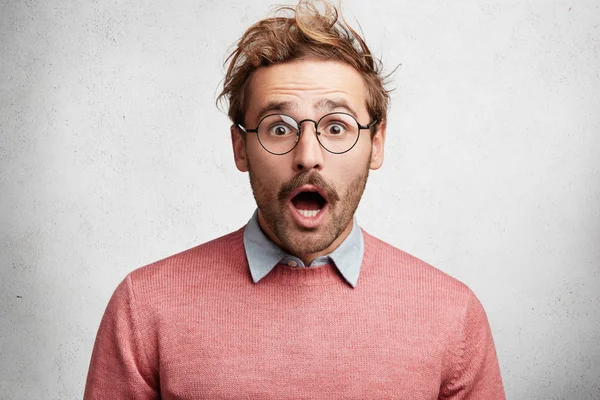 Indoor shot of amazed shocked young man with mustache, stares through round spectacles, being surprised to hear unexpected news, can`t believe eyes, isolated over white concrete background.
