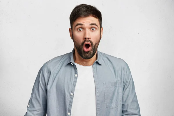 Emotional stunned handsome male has dark hair, mustache and beard, being shocked with sudden raise of prices, expresses great surprise, isolated over white concrete wall. Emotions concept