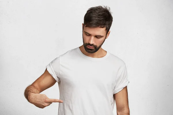 Confident fashionable young bearded male brunet indicates at blank copy space at t shirt, advertises new clothes, area for your promotional text, design or logo. Clothing and people concept.