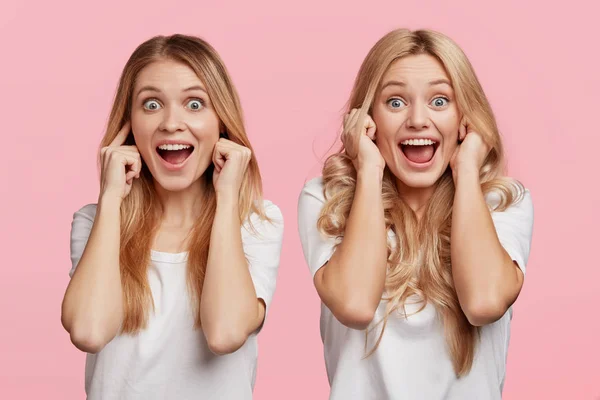 Crazy blonde female plugging ears as hear loud noise, have fun together, pose against pink background. Emotional overjoyed women being on party, don`t want to hear music. Stop this sound, please!