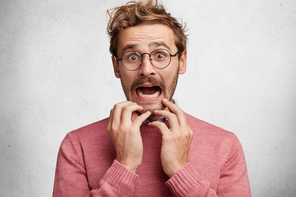 Worried scared fashionable man looks with terrified expression, keeps arms on chin, dressed in pink sweater, isolated over white background. Emotional bearded young guy receives unexpected news