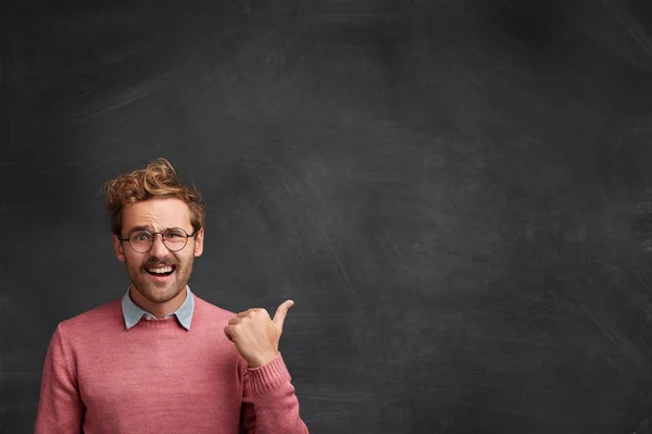 Confident happy handsome young male model dressed in pink sweater with shirt, indicates at blank black chalk copy space for your advertisement or promotional text