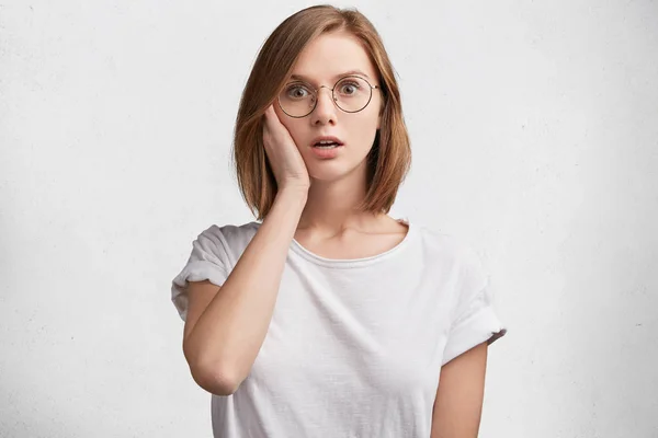 Frightened scared woman wears spectacles, looks with terrified expression, wears white t shirt, afraid of something, expresses negative emotions and feelings, poses against white background.