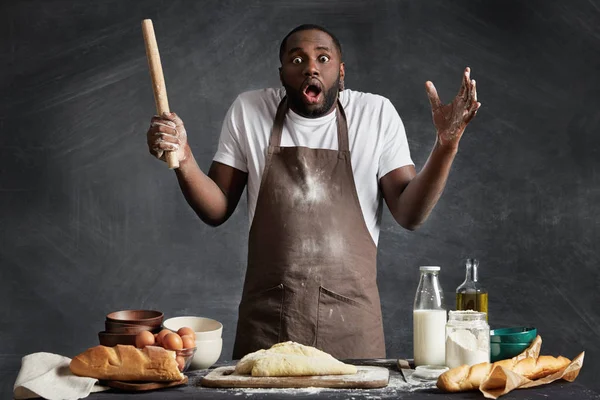 Horrified dark skinned male chef being busy at kitchen, can`t manage bake many cakes for guests, looks surprise at camera, holds rolling pin for making pastry or dough.