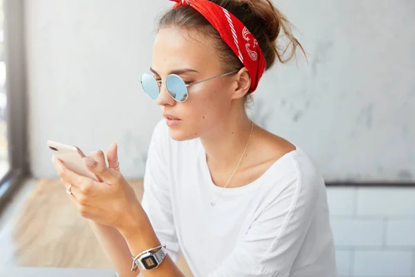 Fashionable pleasant looking female wears red headband, stylish sunglasses, uses smart phone for online communication, texts messages via social networks, uses free internet. People and technology