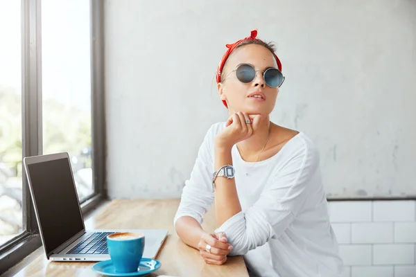 Pensive stylish female wears round sunglasses, white sweater and red bandage, looks aside, works remotely on laptop computer with blank copy space for your advertisement in cozy calm cafeteria.