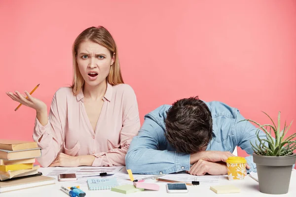 Indignant female model working together with man who asleep, create new scientific research. Exhausted students studying for exams all night on pink background
