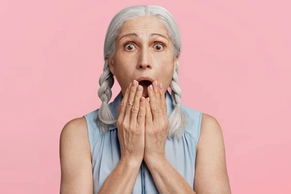 Frightened amazed grey haired female covers mouth, being shocked to hear bad news, expresses great surprise, scared of something, isolated over pink background.