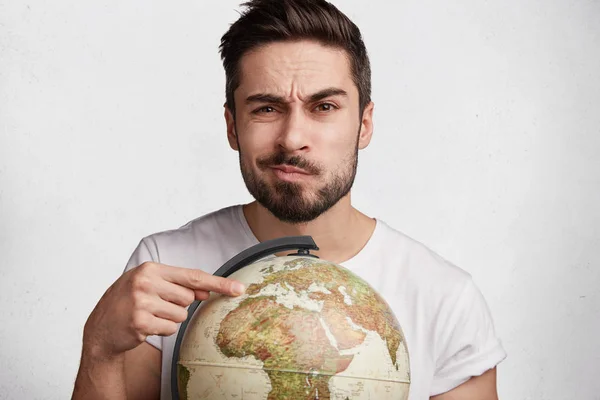 Hesitant male model with stubble indicates at globe, can`t decide where to travel, likes travelling and reaching new destinations, isolated over white background.
