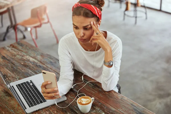 Beautiful young woman enjoys online communication, makes video call via smartphone and earphones, drinks tasty cappuccino, has puzzled expression as has some troubles with internet connection