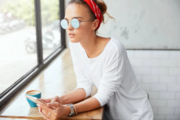 Thoughtful beautiful young female enjoys aroma cappuccino and online communication with friends on smart phone, looks pensively at window. Woman blogger thinks about creating personal website