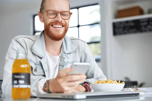 Image of cheerful ginger male with thick beard and mustache wears denim jacket, happy to receive message from best friend on cell phone, has dinner break after work in canteen, smiles happily