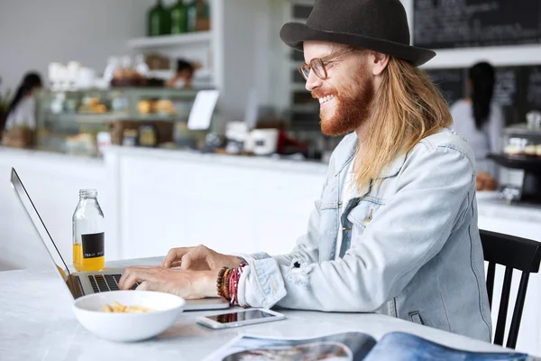 Shot of pleased bearded male in eyewear and hat, dressed in denim jacket, enjoys online communication with girlfriend, share news, keyboards text message, surrounded with modern devices and tasty dish